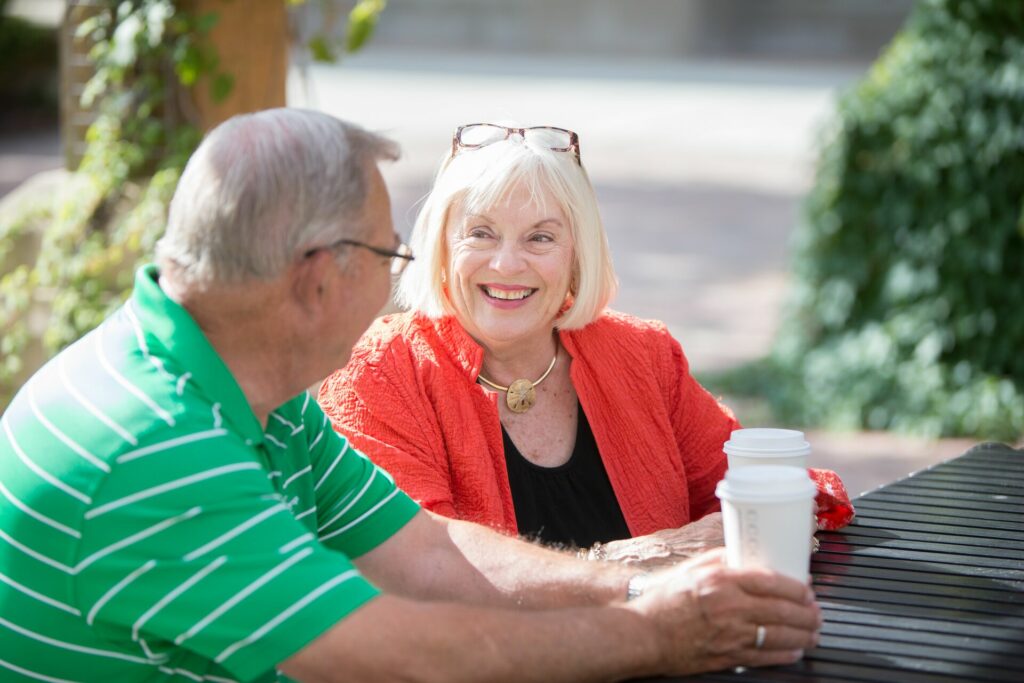 A senior woman smiles at a senior man while sipping out of to go coffee cups outside.
